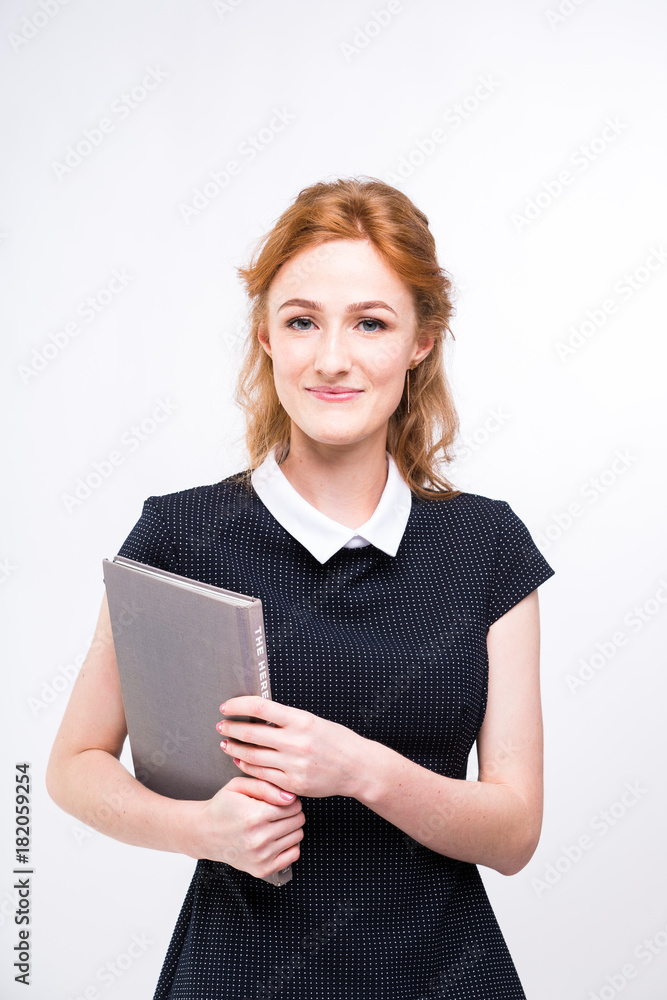 Beautiful girl with red hair and gray book in hands dressed in black dress on white isolated background Beautiful girl with red hair and gray book in hands dressed in black dress on white isolated background