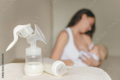 Manual breast pump and bottle with breast milk on the background of mother holding in her hands and breastfeeding baby.