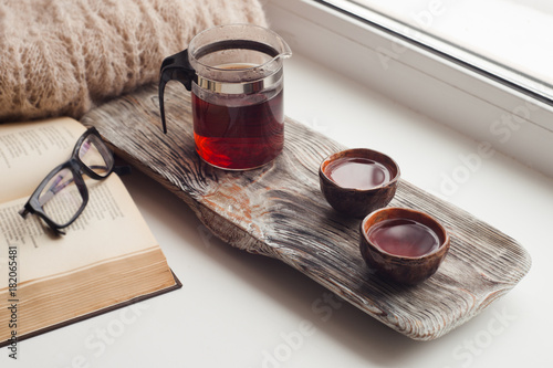 Still life details, cups of asian tea and teapot on vintage wooden tray on windowsill in living room. Lazy winter weekend with a book at home