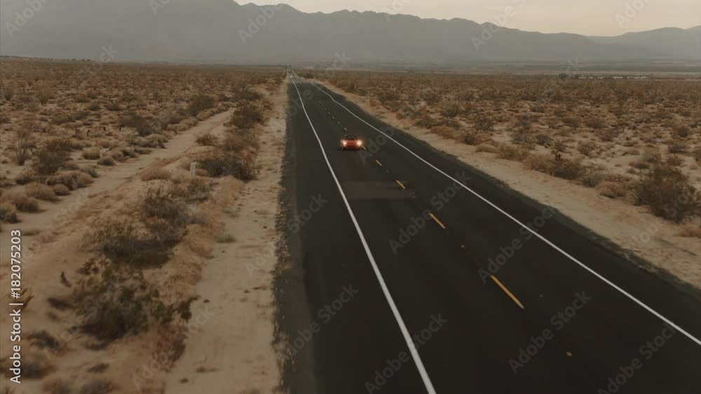 Aerial View Red Classic Car Driving Along Desolate Desert Road At Dusk With Headlights On Aerial View Red Classic Car Driving Along Desolate Desert Road At Dusk With Headlights On