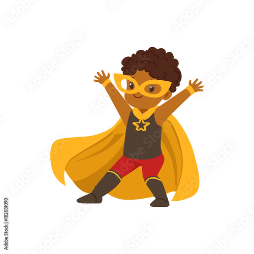 Comic brave black kid in superhero colorful Halloween costume with mask and developing in the wind yellow cape, posing with hands up.