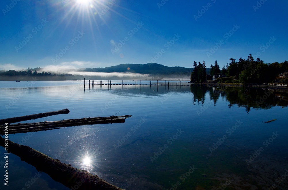 The morning sun rises over Sooke harbour burning off the fog casting its reflection into the calm waters.