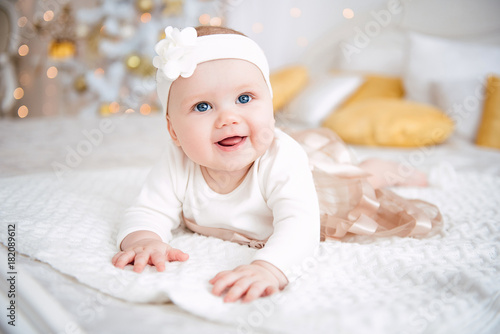 Baby girl wearing cute dress and headband, lies on a white cover in festively decorated room with garland of lights. With surprise watches in the camera, on a background a set of bright fires.