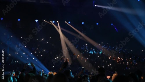 Concert hall with viewers. Audience watching concert on stage in big concert club. Cheering crowd in concert hall. Viewers move in time with the music. Audience is shining with flashlights of phones.