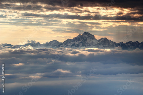 Jagged snowy peaks of Julian Alps range and Triglav, highest mountain of Slovenia, towering dramatically above glowing sea of clouds lit by sun rays light at autumn sunset Triglav National Park Europe © nogreenabove2k