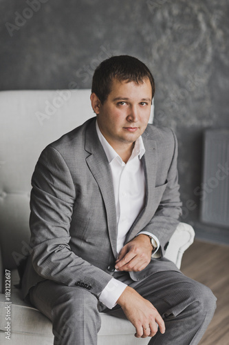 A young man in a gray suit sitting on the sofa on the grey patterned wall 1