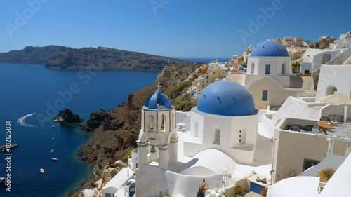 Panning shot of iconic white houses and blue dome churches in Santorini Island, Greece. Sunny day, blue sky