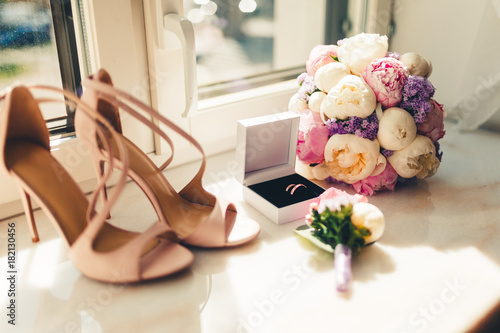 Wedding rings in white box, beige sandals, a bouquet of flowers and boutonniere at the window. Accessories for wedding. The symbols of love, wedding rings,shoes, wedding bouquet. Tonted photo. photo