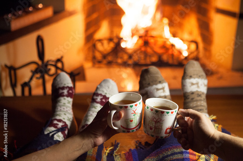 Man and woman in warm knitted socks with cups of hot drink in front of the fireplace