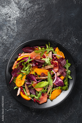 Salad with grilled duck breasts, persimmon, rocca, spinach beet, grapefruit and red onions on black plate on textured anthracite background