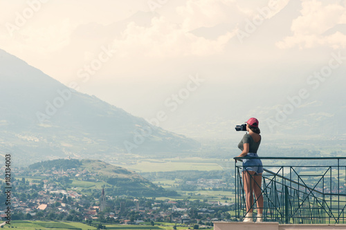 A girl photographer in a red cap with a camera stands on the balcony opposite of Italian mountains and village in South Tirol. Alto Adige