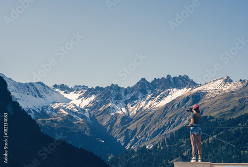 A girl photographer in a red cap with a camera stands on the balcony opposite snow Swiss Alps and forest of national park in Switzerland. Alps of Switzerland on autumn