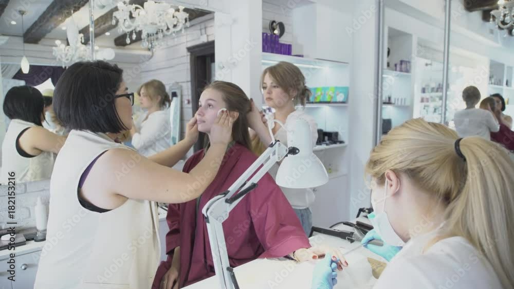 Artist cleanses face of woman and hairdresser does hairstyle indoor. Female cleanses skin of lady with cotton swabs while stylist is engaged in hair and nail master does manicure. Three specialists Artist cleanses face of woman and hairdresser does hairstyle indoor. Female cleanses skin of lady with cotton swabs while stylist is engaged in hair and nail master does manicure. Three specialists