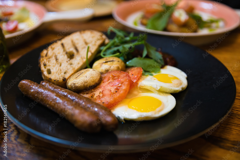 Close focus on dish of american breakfast with grilled pork sausages, fried eggs, toast; mushroom and tomato in Russian restaurant.