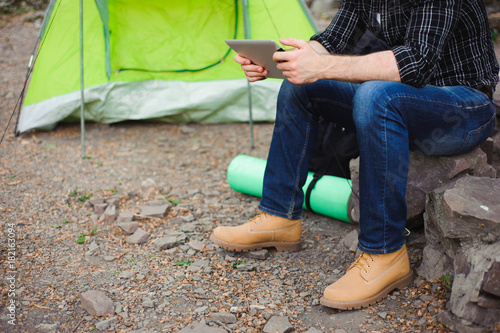 Traveler near a tent with a tablet in the evening. Rocky mountains background.