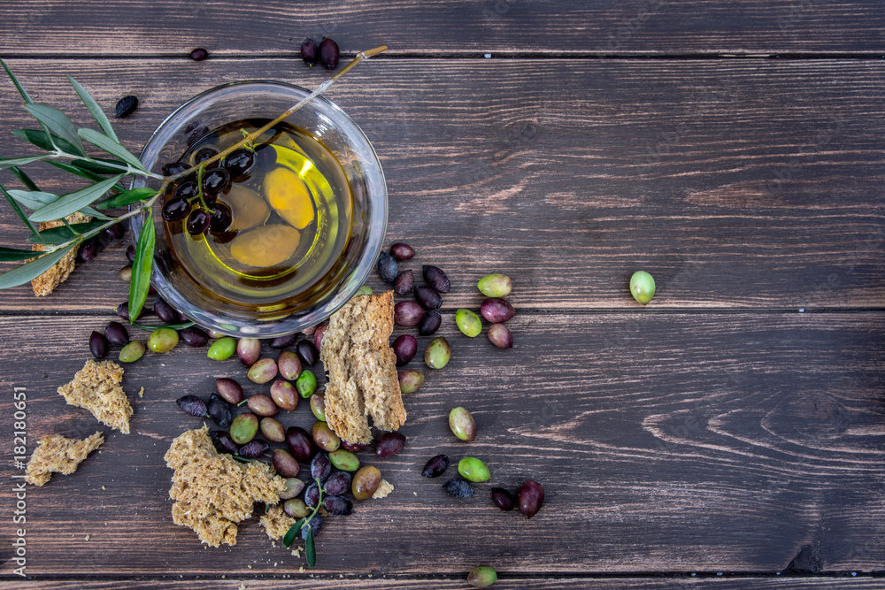 Bowl with extra virgin olive oil, olives, a fresh branch of olive tree and cretan rusk dakos close up on wooden table, Crete, Greece. - obrazy, fototapety, plakaty 