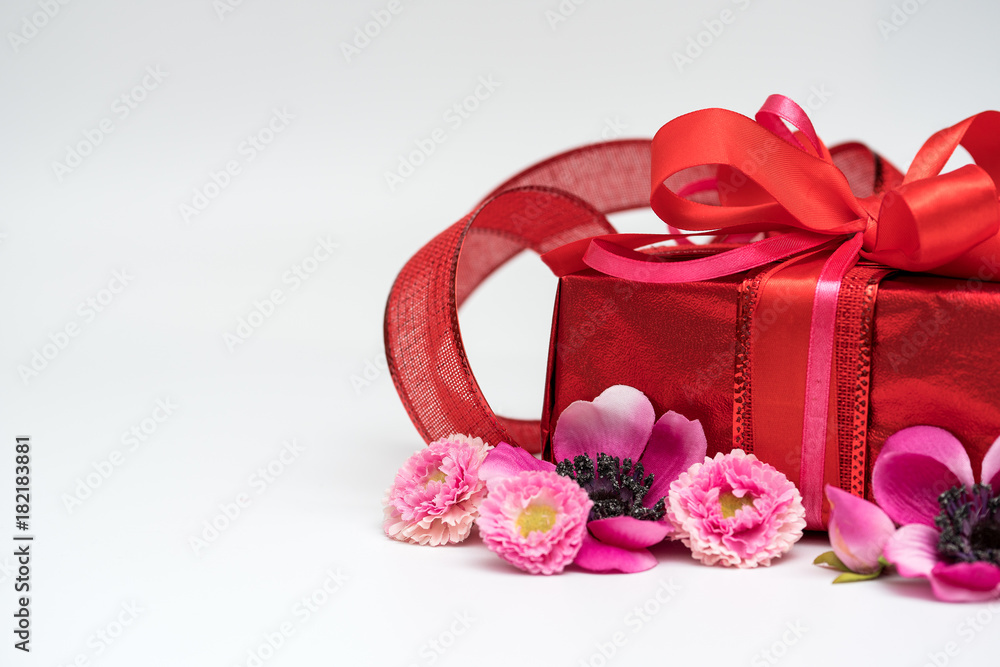 Festive gift box with a bow of red color. Lies on a white background. Next to pink flowers. Festive gift box with a bow of red color. Lies on a white background. Next to pink flowers.