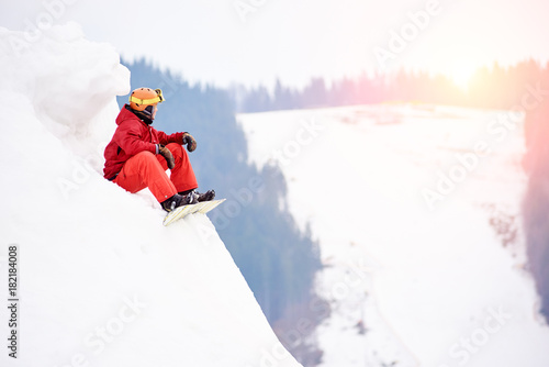 Male snowboarder in a red suit sitting on the top of the snowy hill with snowboard, looking to the sunset. Skiing and snowboarding concept