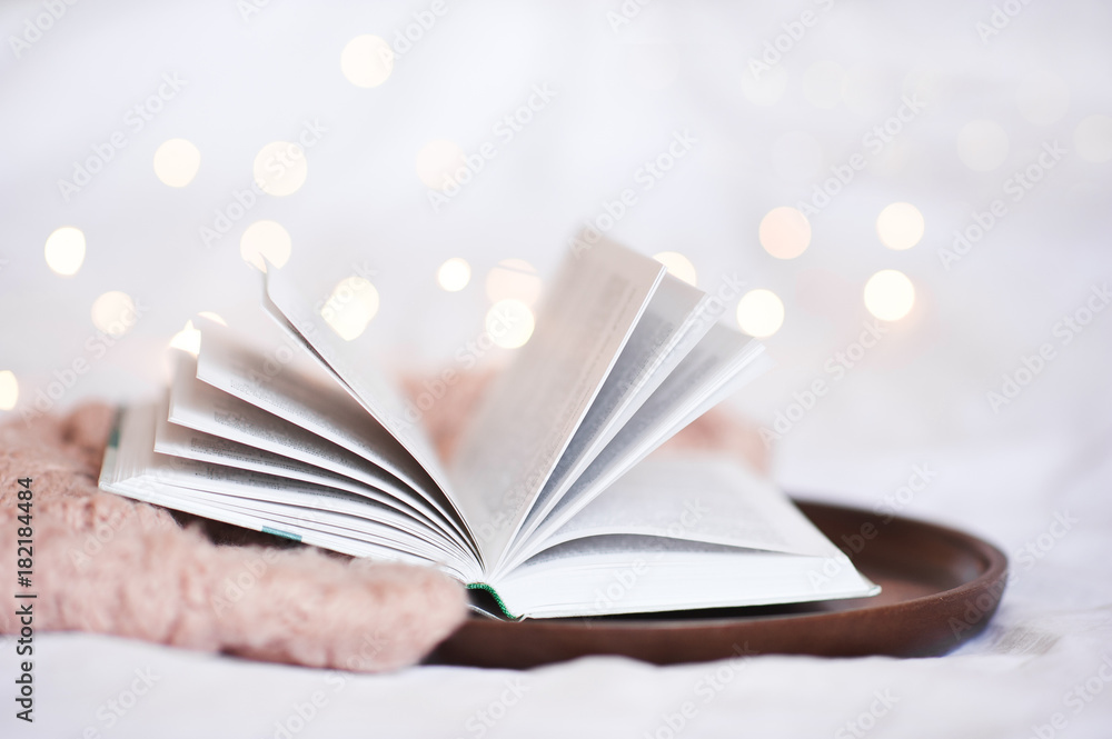 Open book on wooden tray with knitted sweater closeup over Christmas lights. Open book on wooden tray with knitted sweater closeup over Christmas lights.