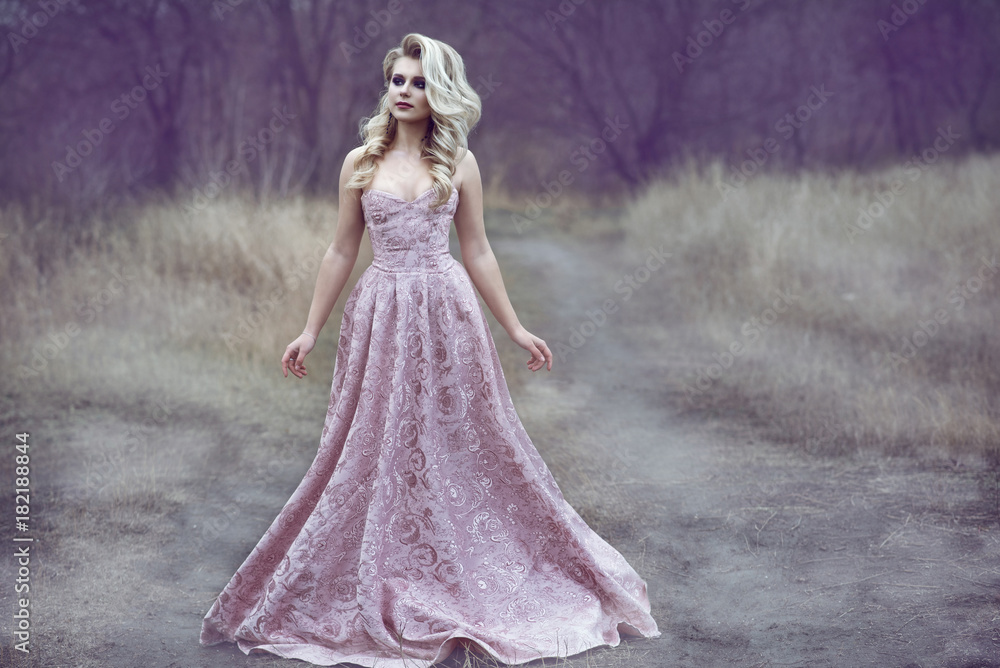 Portrait of gorgeous blond lady with luxuriant hairstyle and beautiful make up wearing luxurious brocade ash rose ball dress walking along the narrow path in the wood. Fairytale concept. Outdoor shot