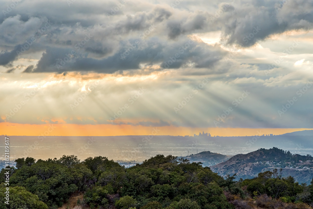 Sunrays pour down through clouds at sunset highlighting the Los Angeles skyline from a distant moutain.