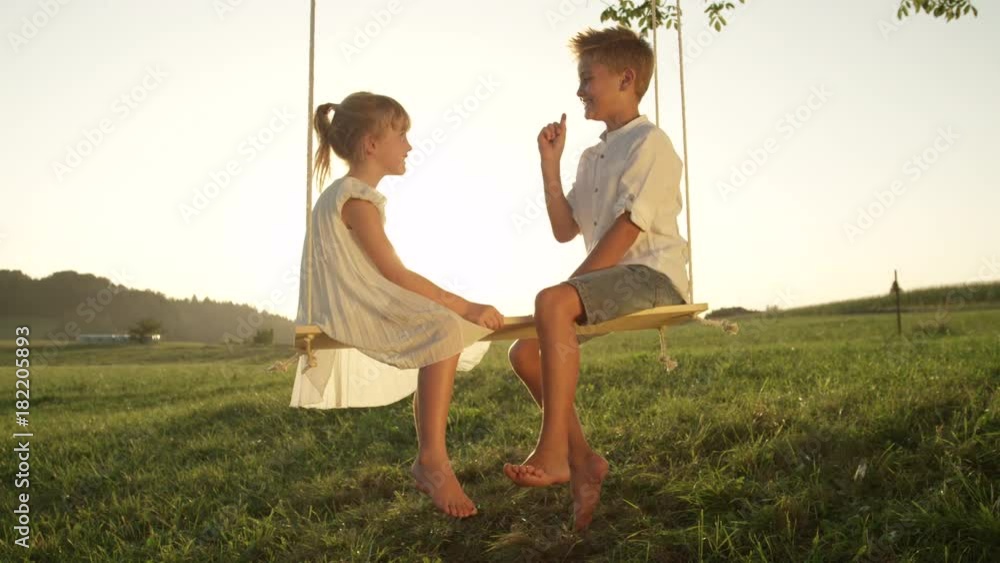 SLOW MOTION CLOSE UP Smiling brother and sister sitting on wooden swing ...