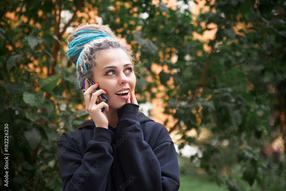 portrait in natural light of a cheerful young girl in hoodie and colored dreadlocks and piercings while talking on a cell phone in a green Park. portrait in natural light of a cheerful young girl in hoodie and colored dreadlocks and piercings while talking on a cell phone in a green Park.