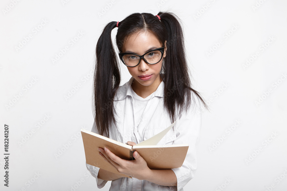 Horizontal studio shot of concentrated college Chinese student girl wearing white medical coat and cat eye glasses holding copybook and writing down results of conducted research or experiment Horizontal studio shot of concentrated college Chinese student girl wearing white medical coat and cat eye glasses holding copybook and writing down results of conducted research or experiment