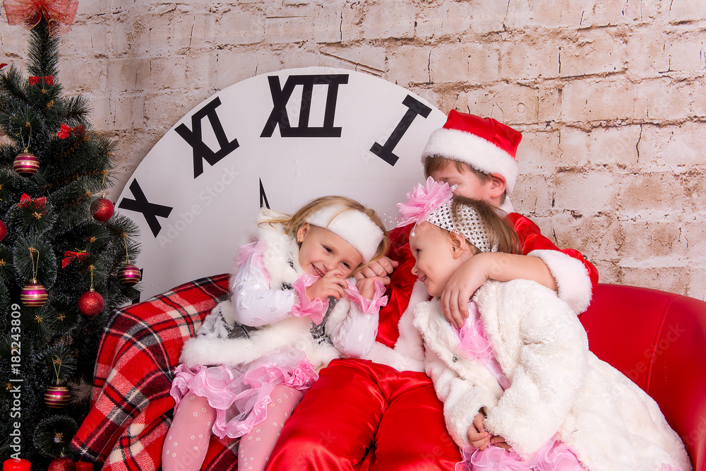 Obraz premium The children, a boy dressed as Santa Claus and two girls in white coats posing against the backdrop of the Christmas decorations in the studio.