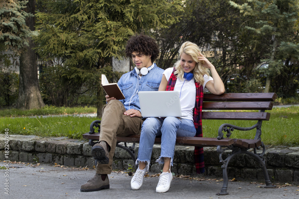 Young couple sitting on bench in park. He is reading a book and she is typing on laptop