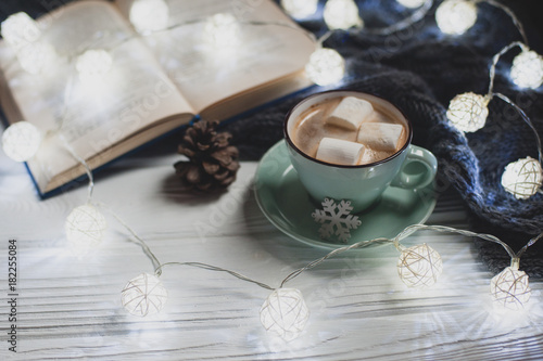 Cozy winter home. Cup of cocoa with marshmallows, warm knitted sweater, open book, Christmas garland, lump on a white wooden table. Atmosphere of a pleasant evening for reading.