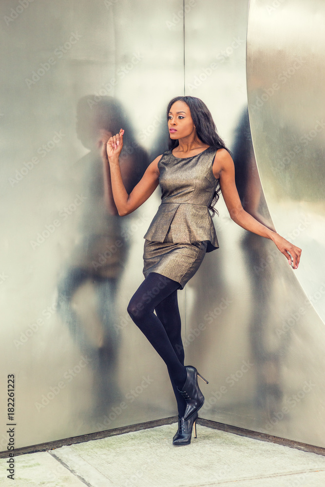 African American Business Woman Fashion in New York. Lady wearing metal  crystal sleeveless mini dress top, skit suit, black leggings, high heels,  stands against silver metal wall, looking forward.. Stock Photo