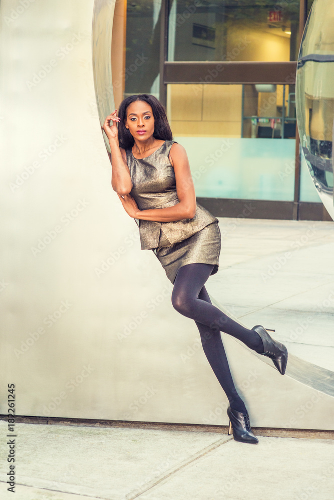 African American Business Woman Fashion in New York. Lady wearing metal  crystal sleeveless mini dress top, skit suit, black leggings, high heels,  leans against silver metal wall, relaxing, thinking.. Stock Photo