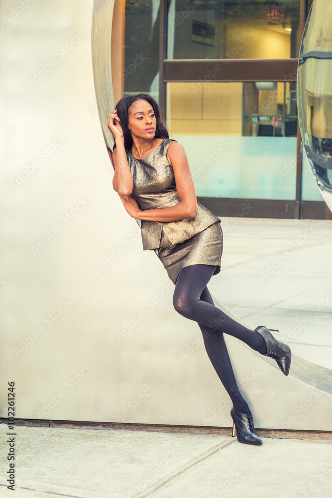 African American Business Woman Fashion in New York. Lady wearing metal  crystal sleeveless mini dress top, skit suit, black leggings, high heels,  leans against silver metal wall, relaxing, thinking.. Stock-Foto | Adobe