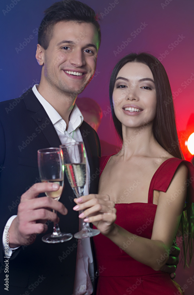 A group of young cheerful and happy friends are dancing at a party. A man and a woman are dancing in the foreground and clinking glasses with champagne A group of young cheerful and happy friends are dancing at a party. A man and a woman are dancing in the foreground and clinking glasses with champagne