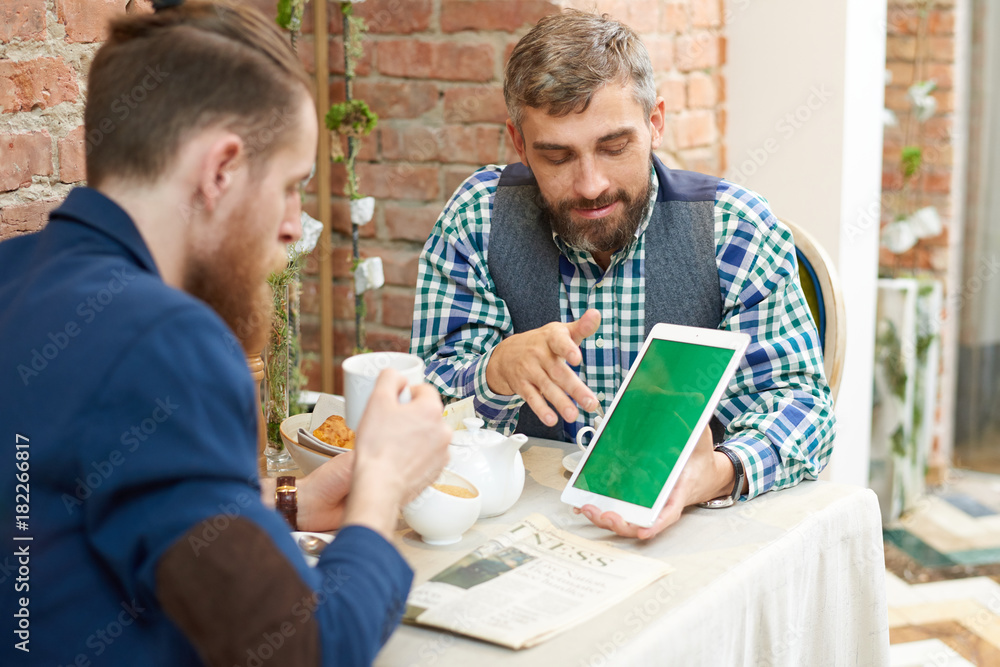 Bearded middle-aged man wearing checked shirt and waistcoat showing statistic data to his male colleague with help of digital tablet while having informal working meeting at cafe.
