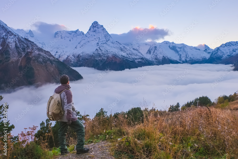 Landscape in the high mountains of the Caucasus after sunset with a guy tourist standing on the edge of a cliff and looking at a valley hidden by clouds