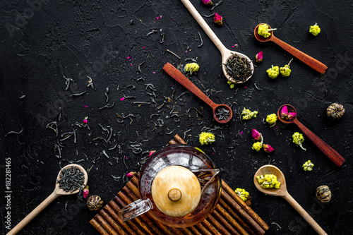 Brew the aromatic tea. Tea pot near wooden spoons with dried tea leaves, flowers and spices on black wooden background top view copyspace