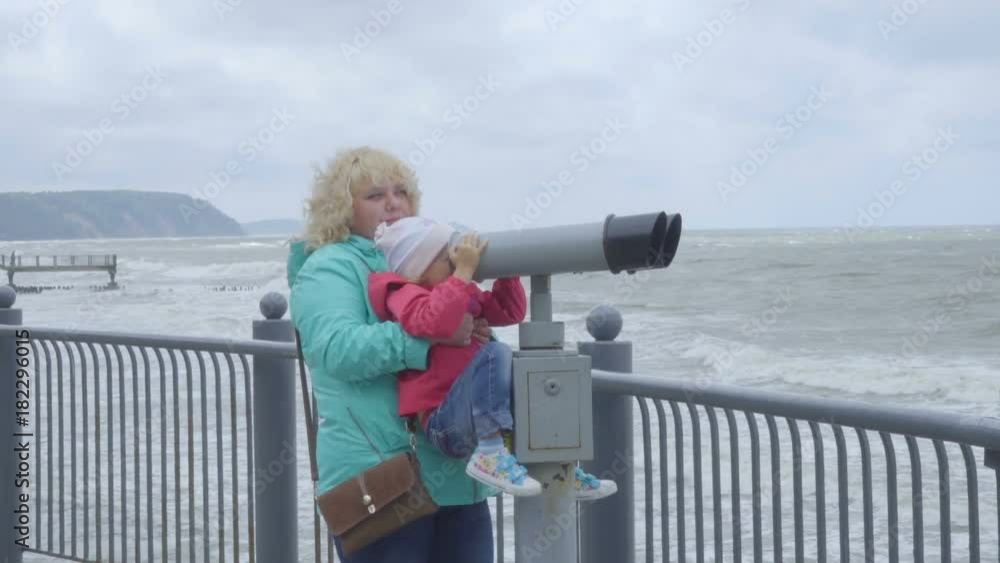 Travel and tourism. Woman and little girl looking through tourist binoculars at the sea or ocean shore. Mother and child look through binoculars on storm of sea. Fat woman holds daughter in her arms. Travel and tourism. Woman and little girl looking through tourist binoculars at the sea or ocean shore. Mother and child look through binoculars on storm of sea. Fat woman holds daughter in her arms.