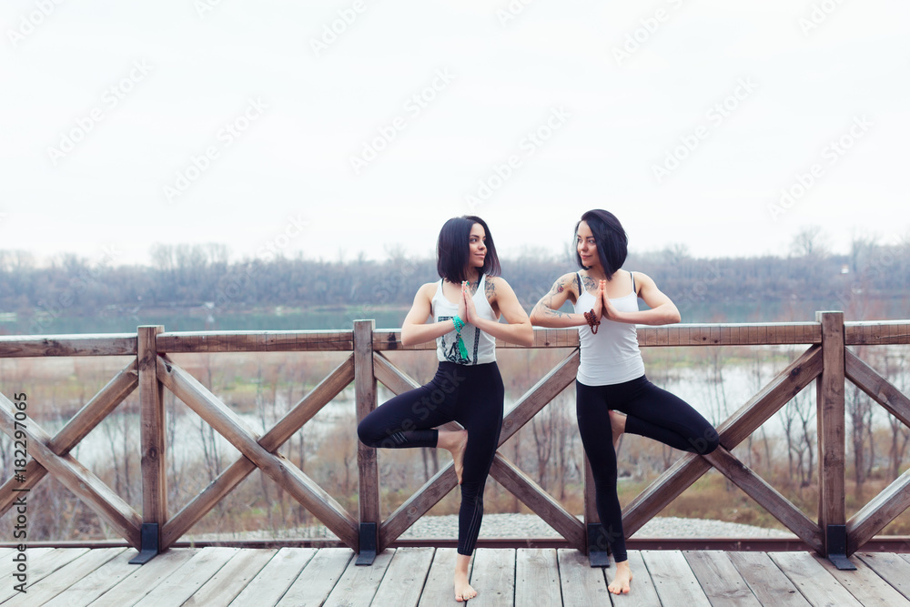 Two attractive sport women twins work out tree yoga pose on black mat outdoors on nature. Group of young women stretching on wooden bridge on spring park