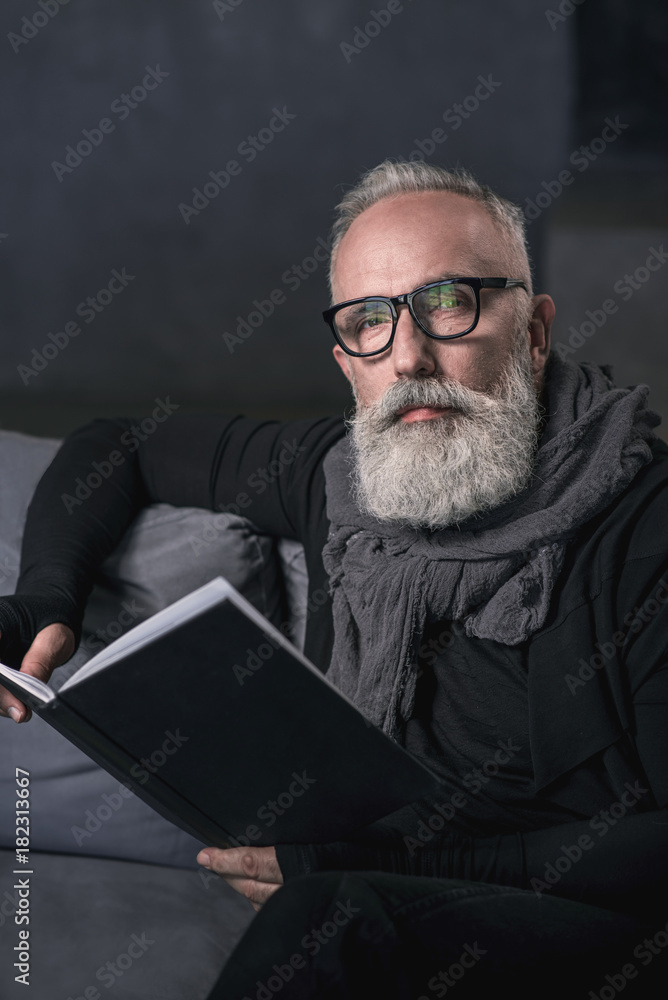 Portrait of serious bearded grandfather reading interesting book while situating on cozy sofa in apartment. Leisure concept Portrait of serious bearded grandfather reading interesting book while situating on cozy sofa in apartment. Leisure concept