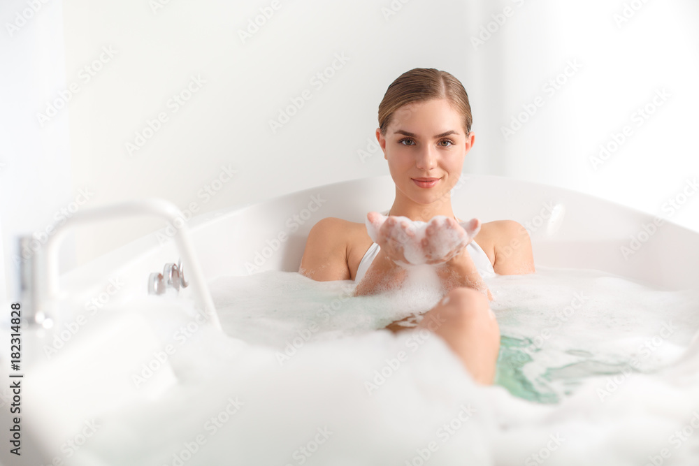 Portrait of confident girl lying in bathtub and keeping water bubbles in arms. She is looking at camera and smiling Portrait of confident girl lying in bathtub and keeping water bubbles in arms. She is looking at camera and smiling