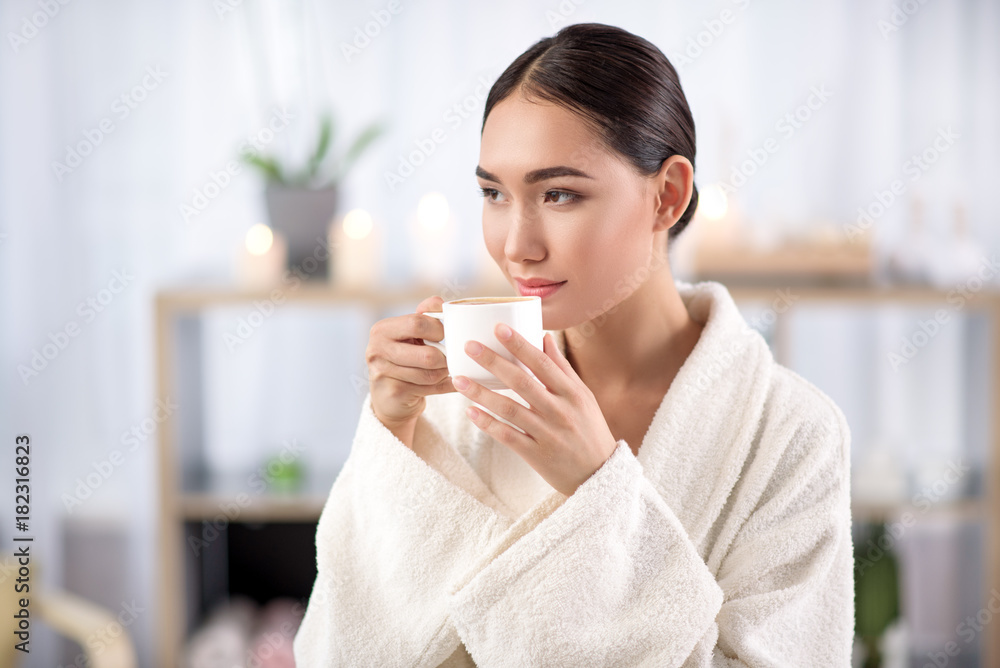 Fresh espresso. Gorgeous asian girl is drinking hot coffee while resting in spa salon. She is sitting in white terry bathrobe while looking aside with slight smile. Shelves with candles in background Fresh espresso. Gorgeous asian girl is drinking hot coffee while resting in spa salon. She is sitting in white terry bathrobe while looking aside with slight smile. Shelves with candles in background