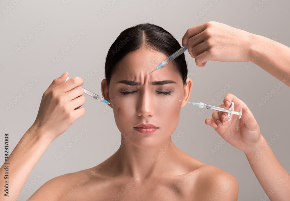 Obraz premium Portrait of naked young asian woman is standing and closing her eyes in pain while getting prick of botox injection in her cheekbones and brow area. Girl is frowning her forehead. Isolated background
