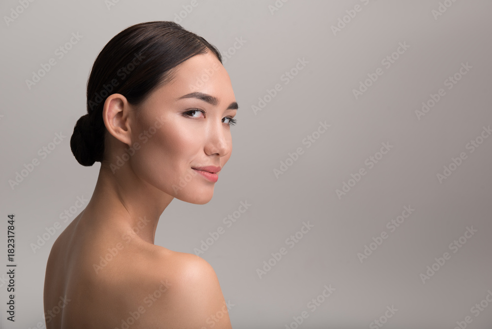 Foto Stock Fresh skin concept. Side view of elegant naked young asian woman  is looking at camera with slight smile while demonstrating her perfect body.  Isolated background with copy space in the