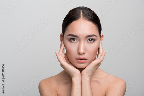 Natural beauty concept. Portrait of attractive dark-haired young asian woman is touching her skin while posing naked. She is looking at camera confidently. Isolated background