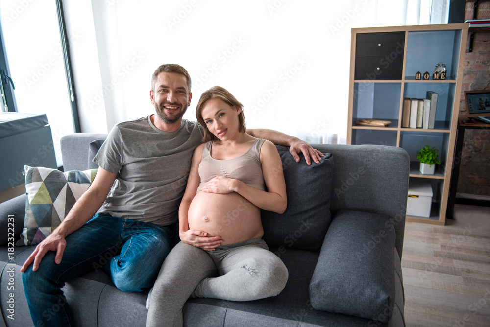 Full length portrait of joyful young family relaxing at home together. Man is embracing expectant woman and smiling while sitting on couch