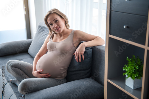 Portrait of joyful expectant mother is sitting on comfortable couch with relaxation. She is looking at camera and laughing