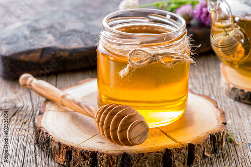 jar of honey with honey dipper on  wooden slice close-up  with teapot and flowers
 photo