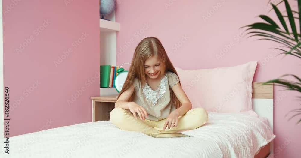 Girl 9 age using tablet pc and sitting on the bed and smiling at camera Girl 9 age using tablet pc and sitting on the bed and smiling at camera
